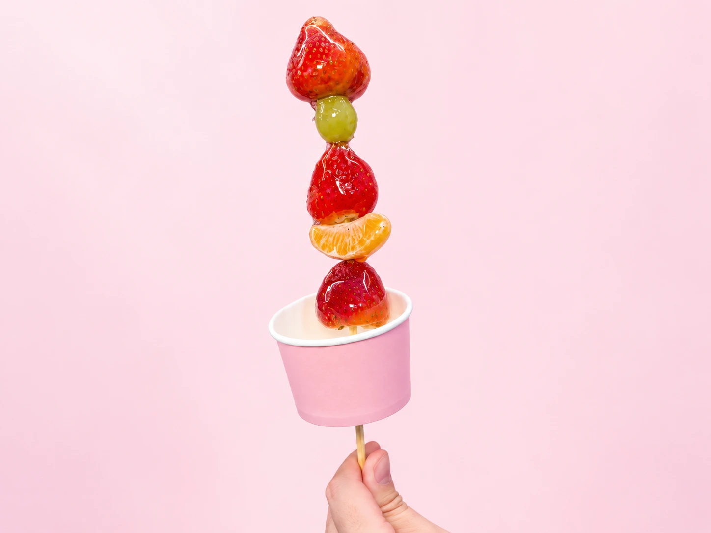 Hand-held TE’AMO strawberry, grape and orange tanghulu in a pink paper cup